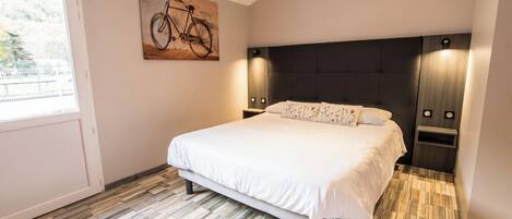 A bedroom with a large bed, a headboard with integrated lighting, a bicycle artwork on the wall, and a window with a view of greenery.