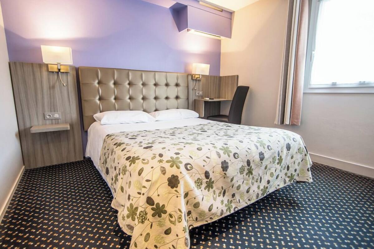 A hotel room with a tufted headboard, a bed with a floral bedspread, a small bedside table, and a chair.