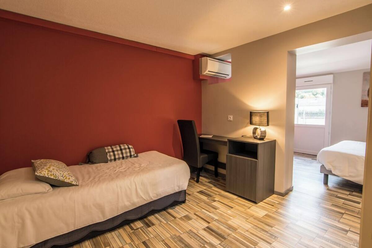 A compact bedroom with a bed, desk, and chair, an air conditioner, and a view through an open door.