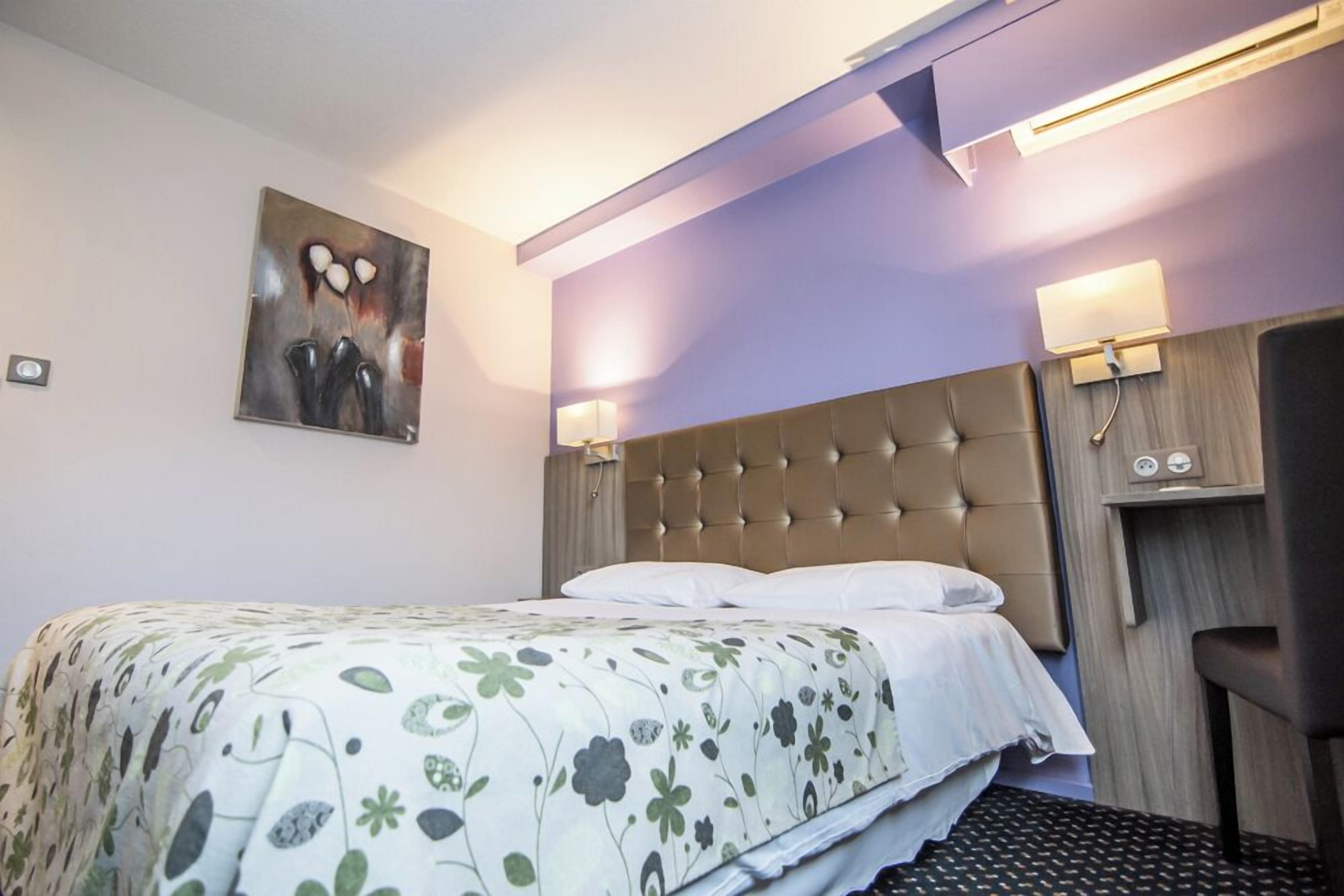 A hotel room with a tufted headboard, a floral bedspread, a painting on the wall, and two wall-mounted lamps.