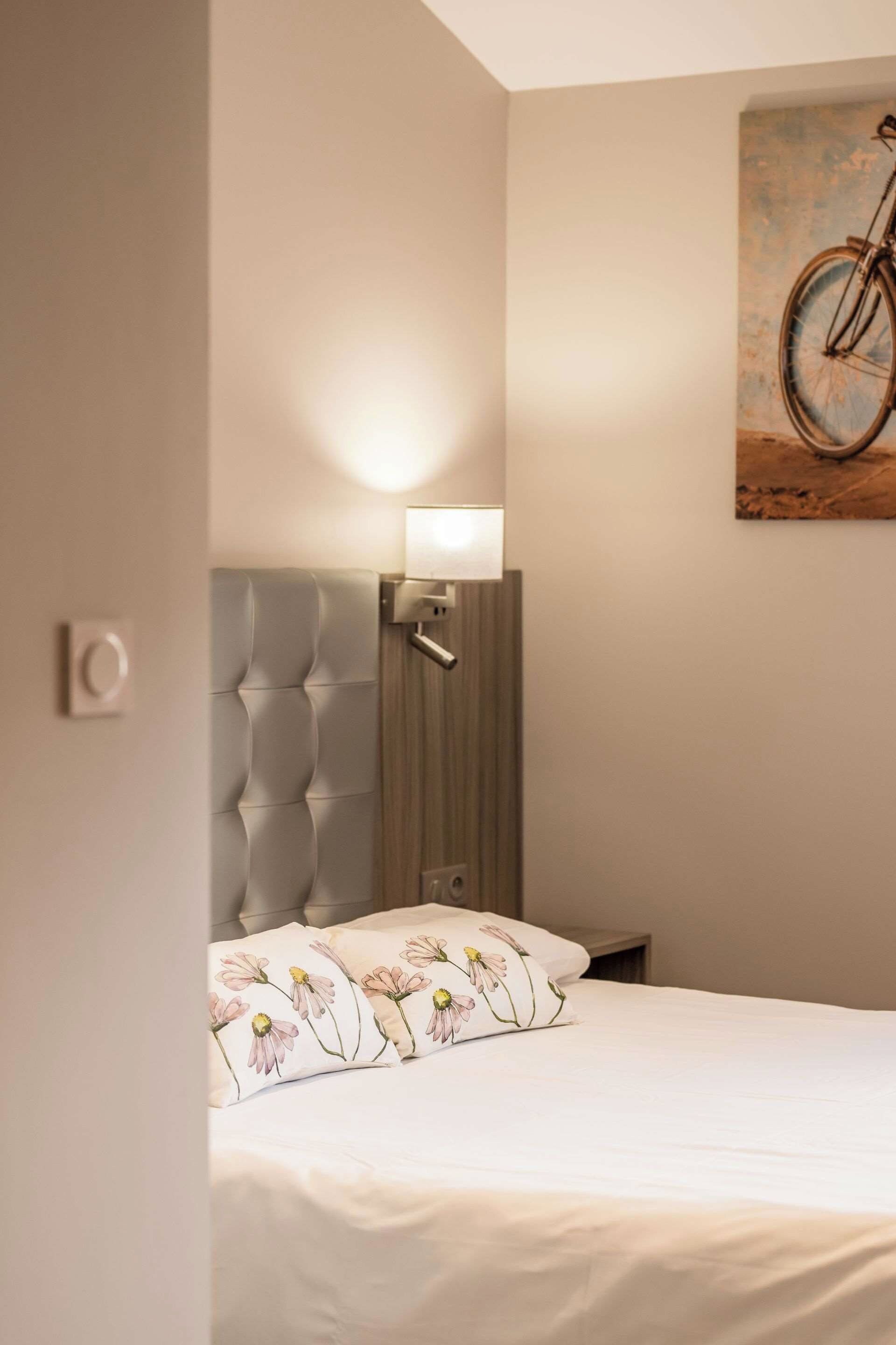 A bedroom with a bed, a nightstand, a wall-mounted lamp, and a painting of a bicycle.