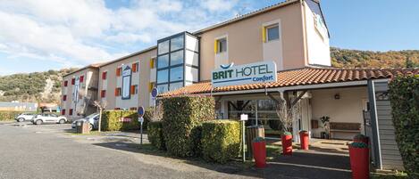 Een hotel met een bord waarop 'BRIT HOTEL Comfort' staat en een pannendak. Er staan auto's geparkeerd en de lucht is helder.