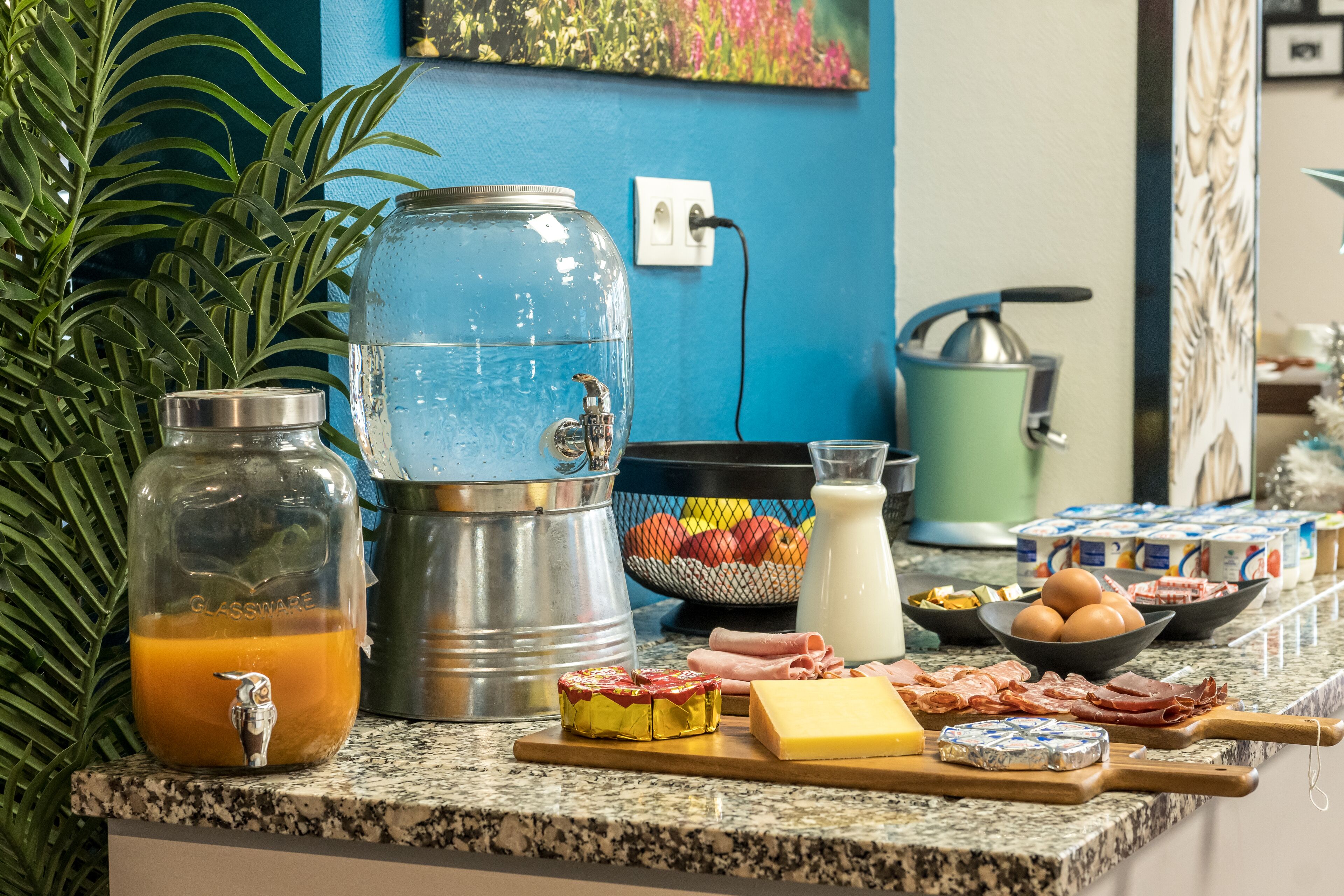 Una encimera de cocina con dispensador de agua, cafetera y diversos alimentos como queso, jamón y huevos.