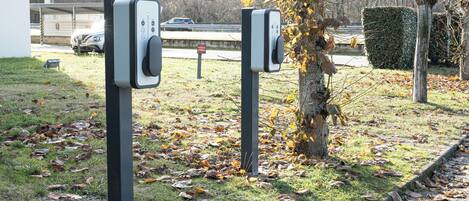 Dos estaciones de carga para vehículos eléctricos en un área con césped, cerca de un árbol y un seto.