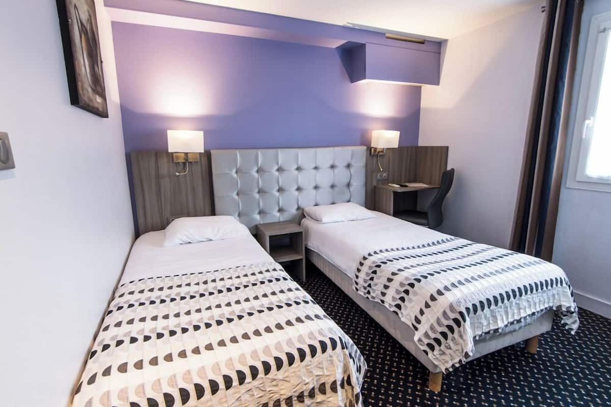 A hotel room with two beds, a headboard, bedside lamps, a small table, and a window with curtains.