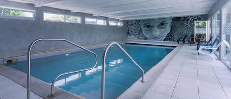 Una piscina interior con agua cristalina, barandillas de metal y un mural en la pared.