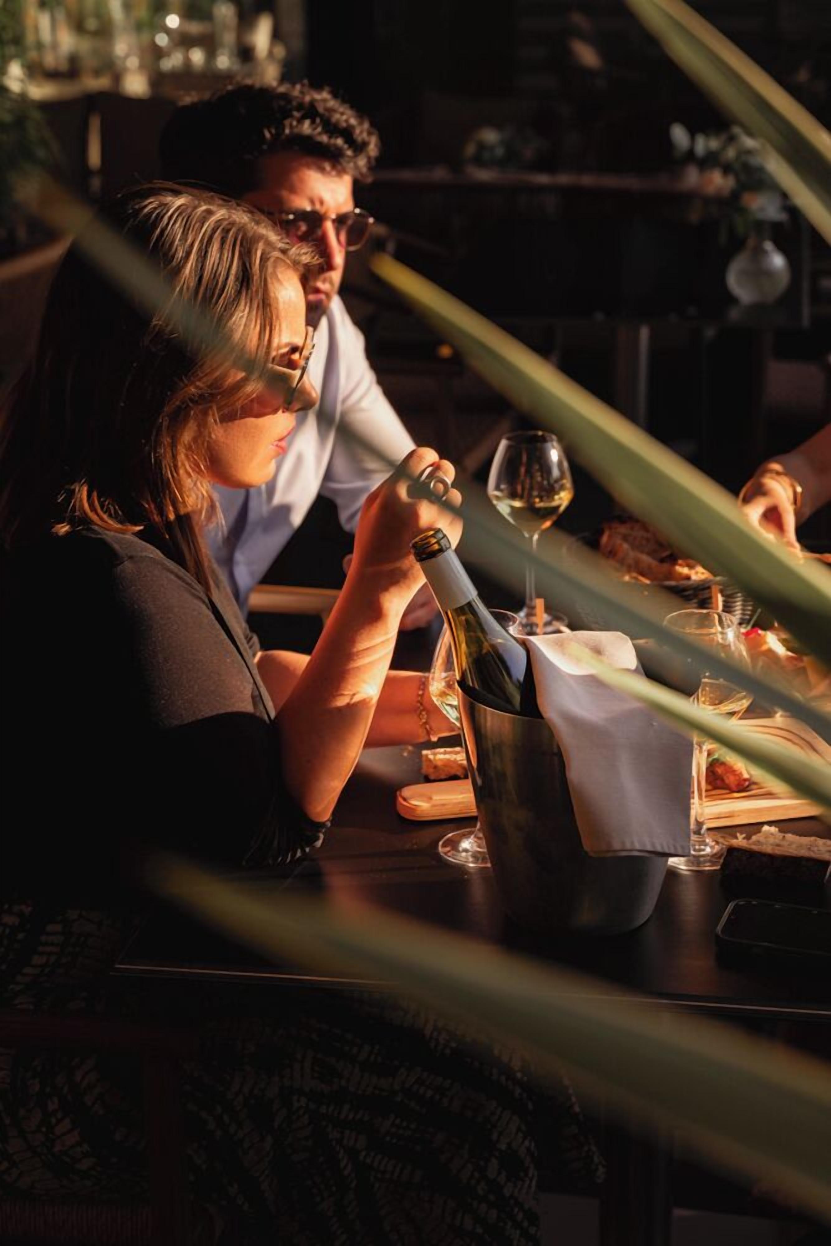 Un couple est assis à une table dans un restaurant, avec une bouteille de vin et un verre devant eux.