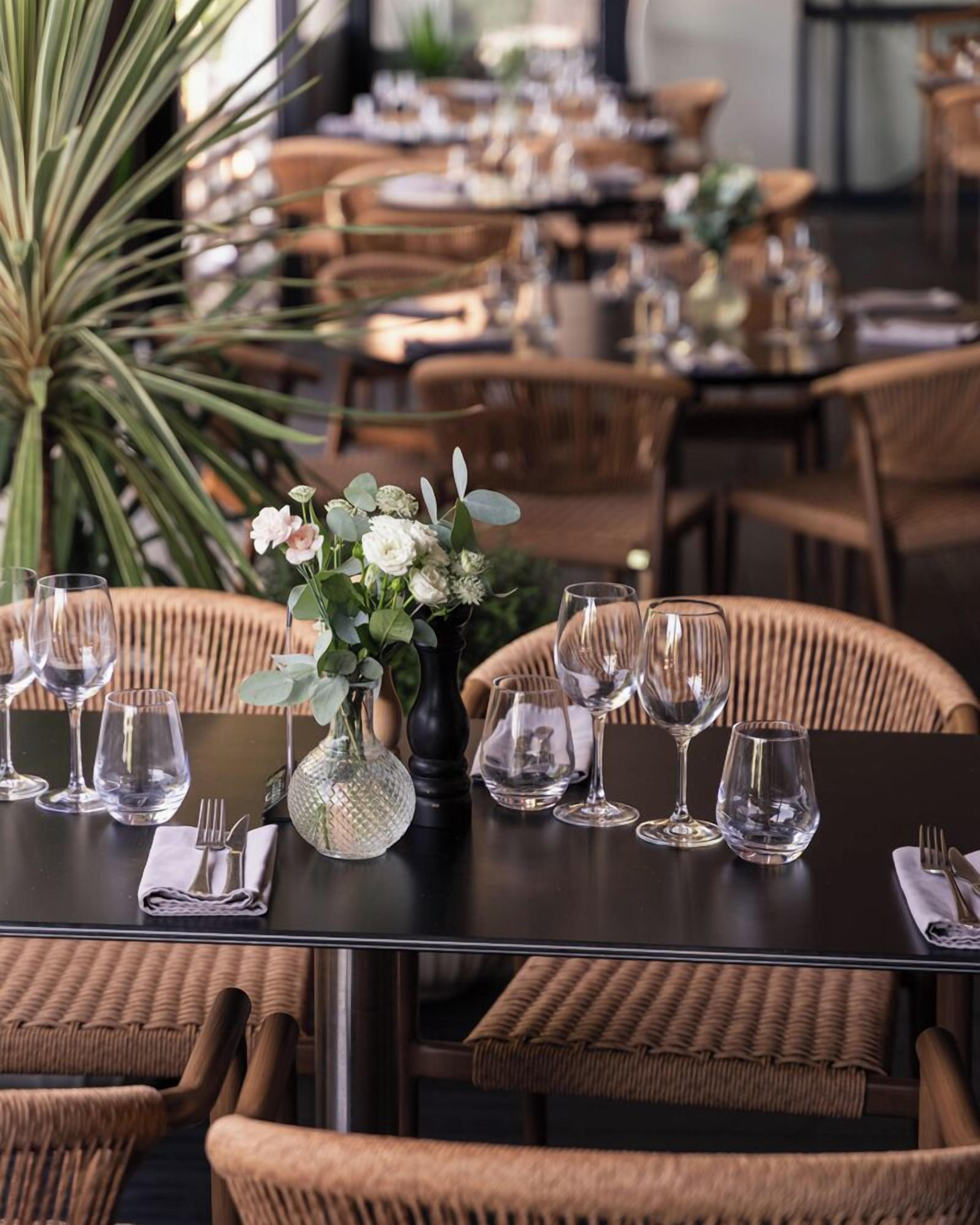 A dining area with wicker chairs and tables, each set with wine glasses and cutlery, and a centerpiece of flowers.