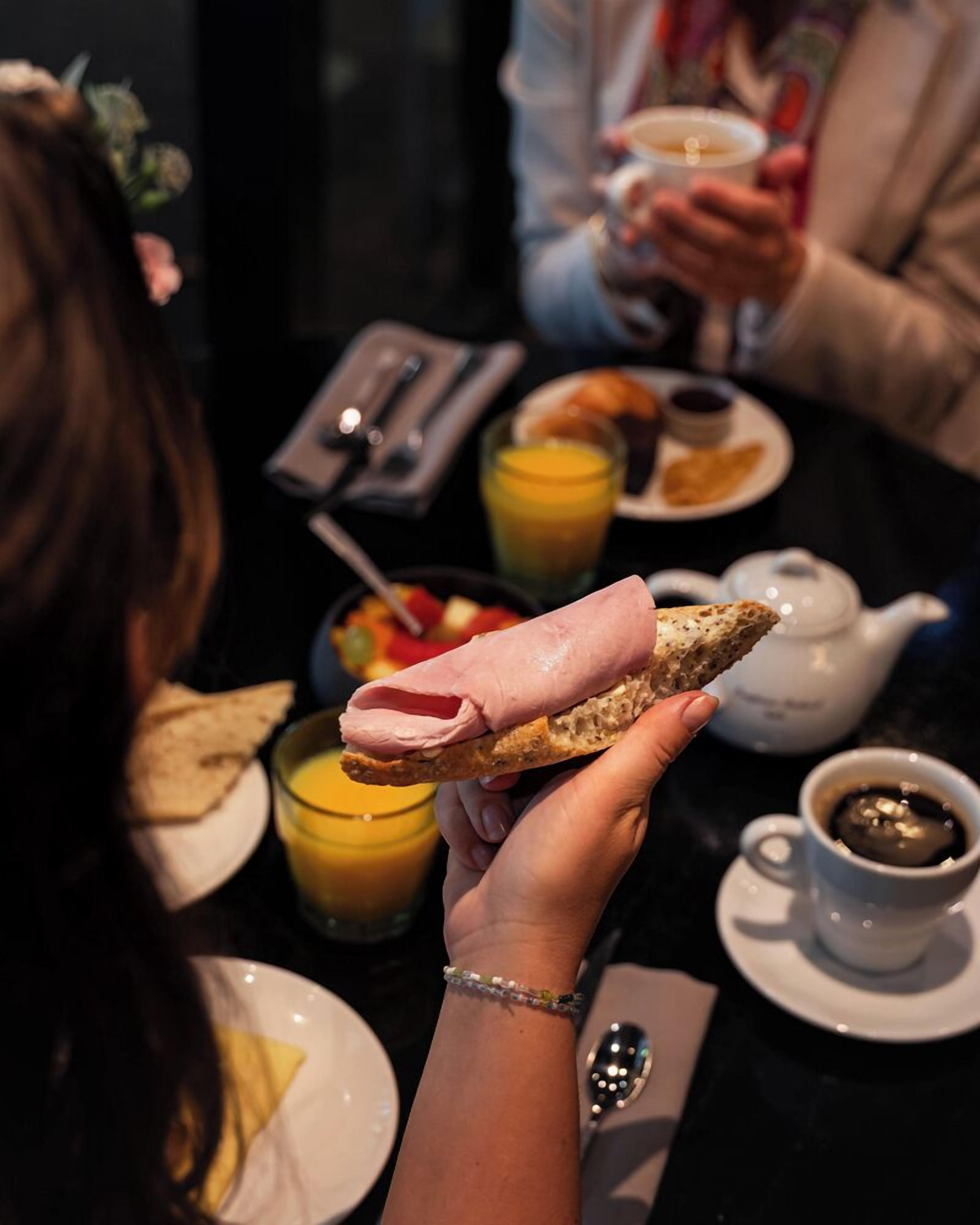 A person holding a sandwich with a slice of ham and cheese, a glass of orange juice, and a cup of coffee on a table.