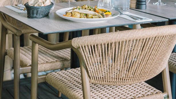 Una mesa de comedor preparada con platos, vasos y una cesta de pan, rodeada de sillas de mimbre.