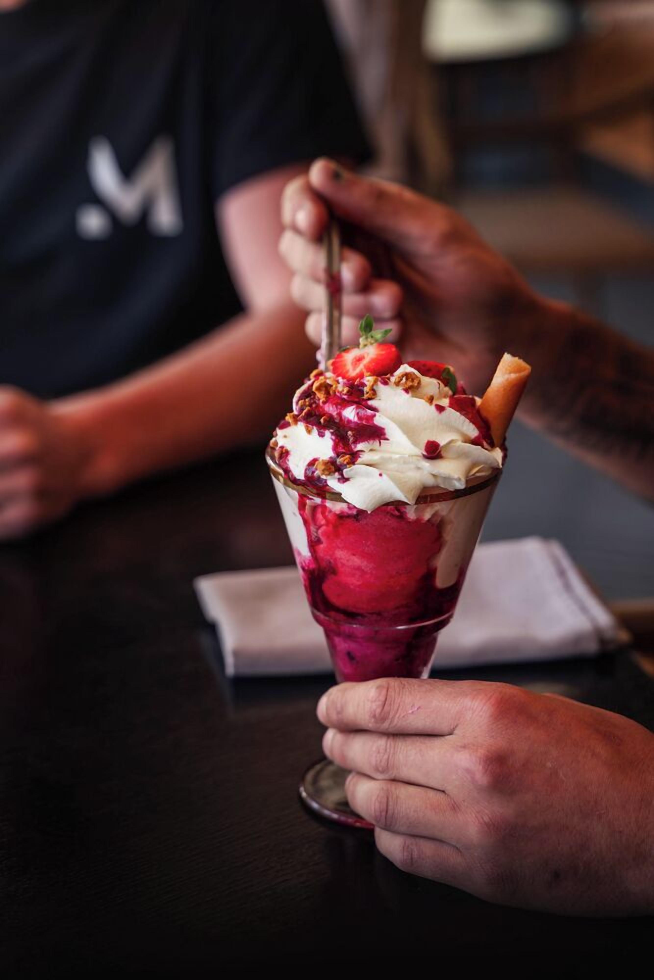 A person holding a dessert with a waffle cone, topped with whipped cream, strawberries, and granola.
