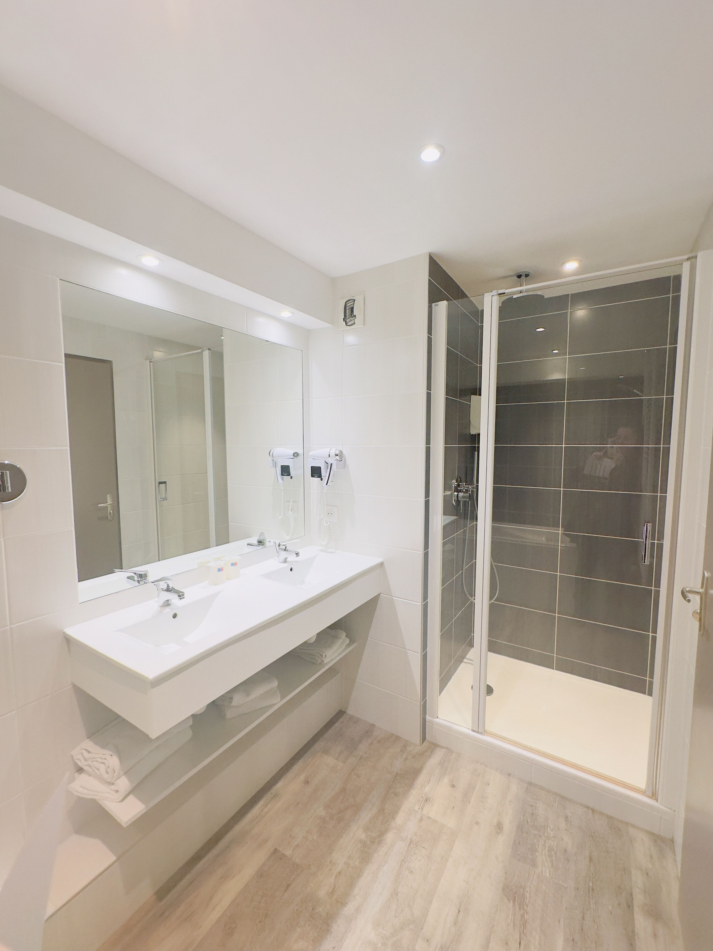 A modern bathroom with a large mirror, two sinks, a shower enclosure, and a wooden floor.
