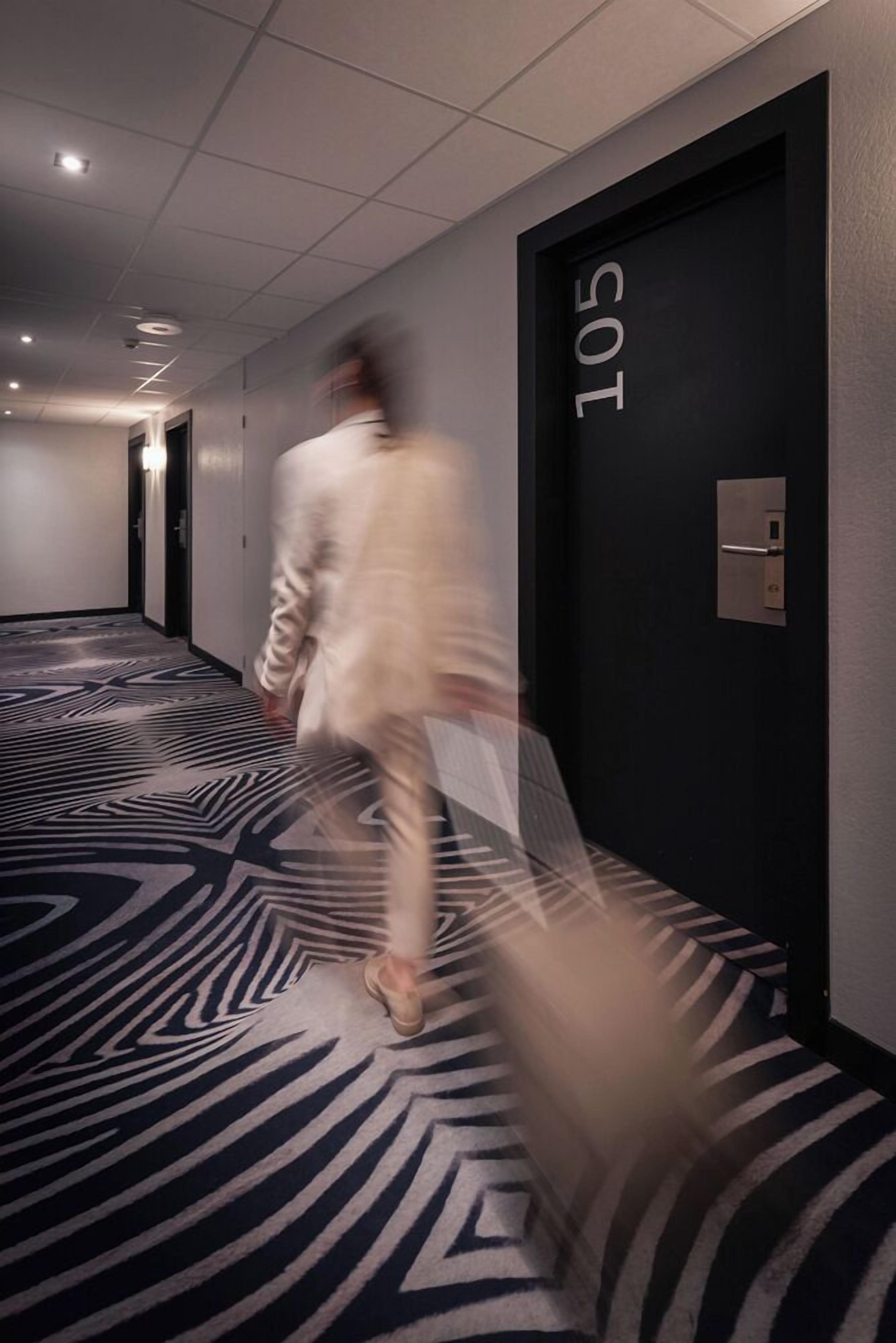 A corridor with a patterned carpet, a door labeled "105", and a motion-blurred person walking.