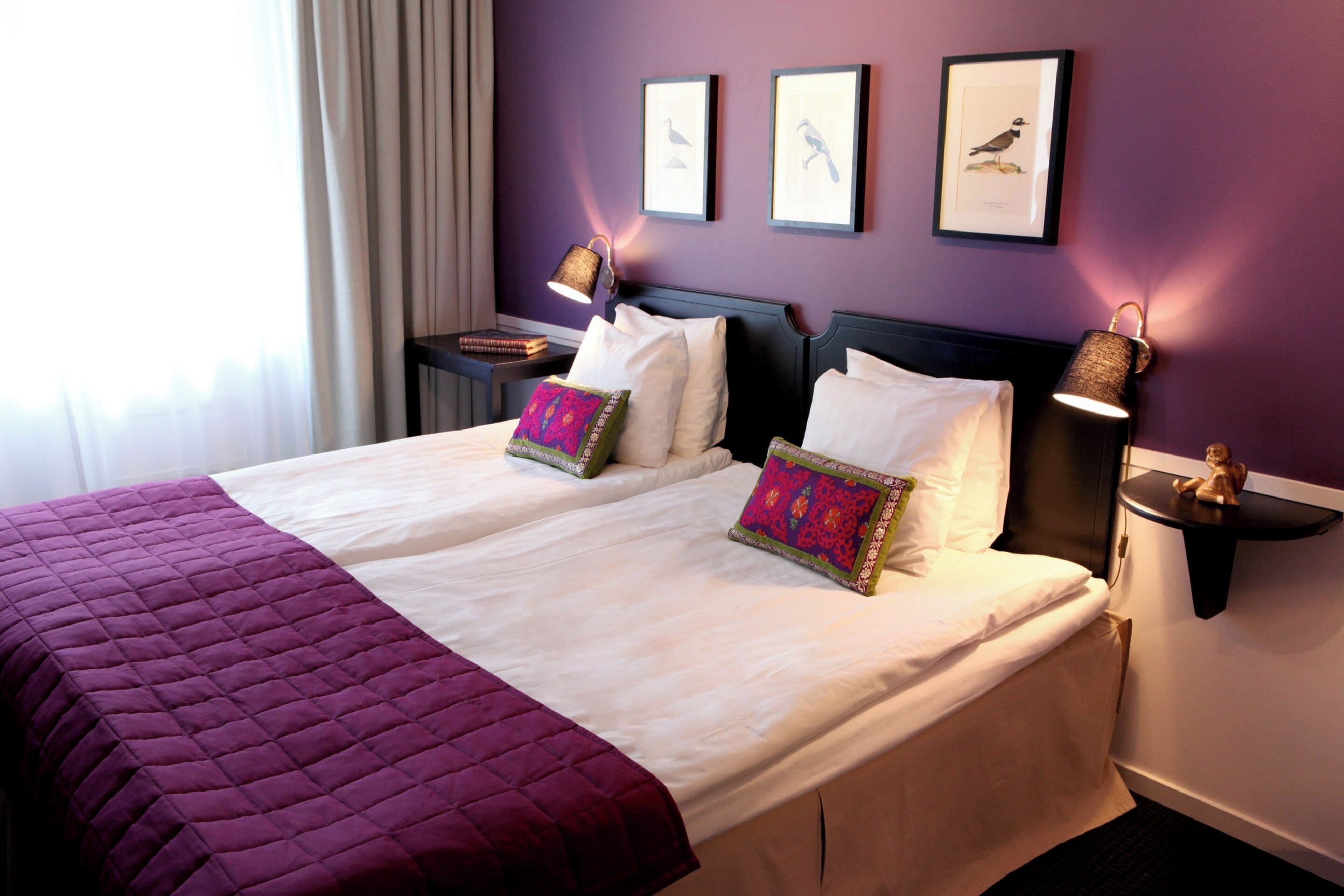 A double bed with a purple quilt, two bedside tables with lamps, and framed bird pictures on the wall.