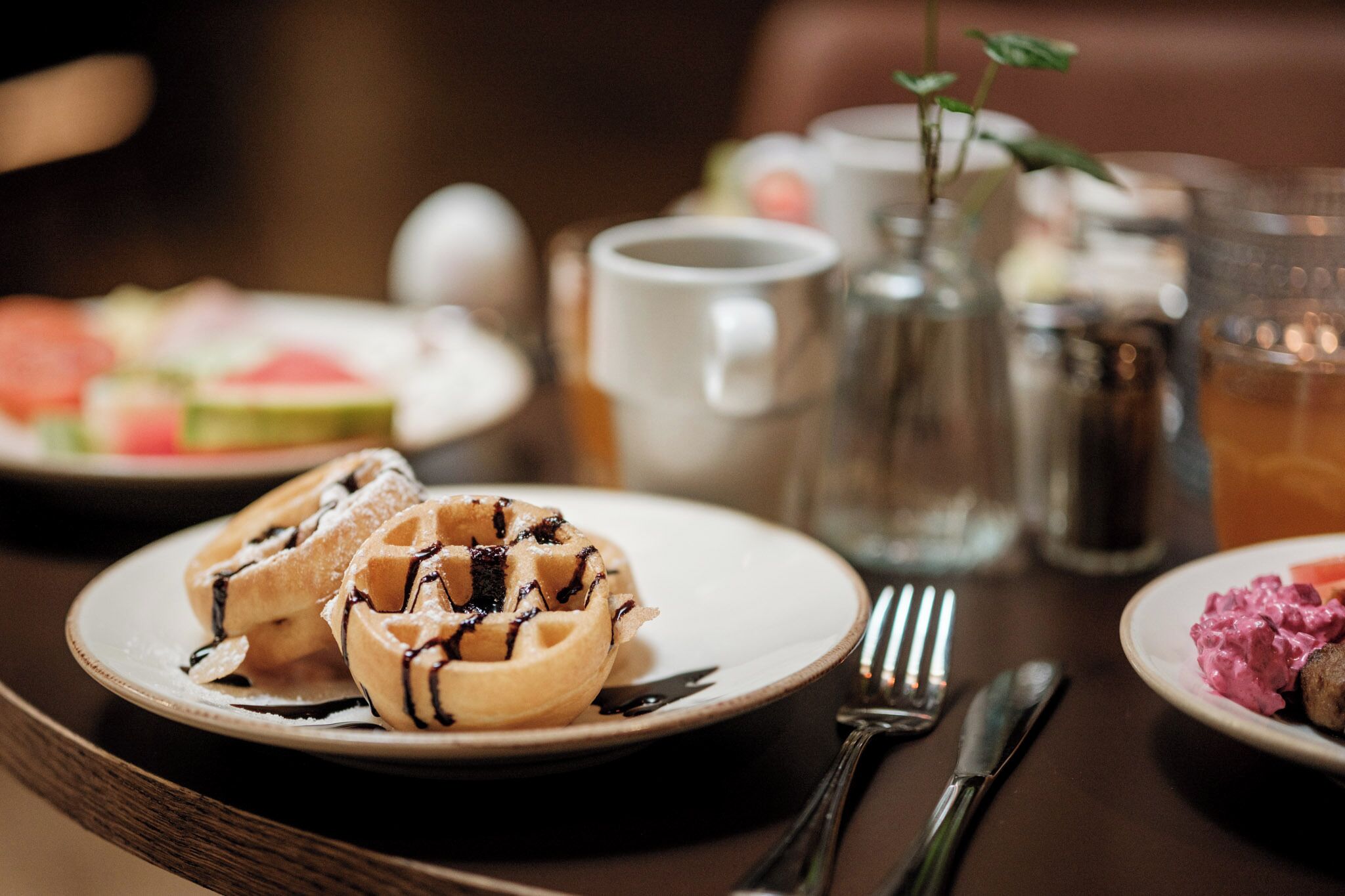 A dining table with a plate of waffles, a pitcher, a mug, and a glass of tea.