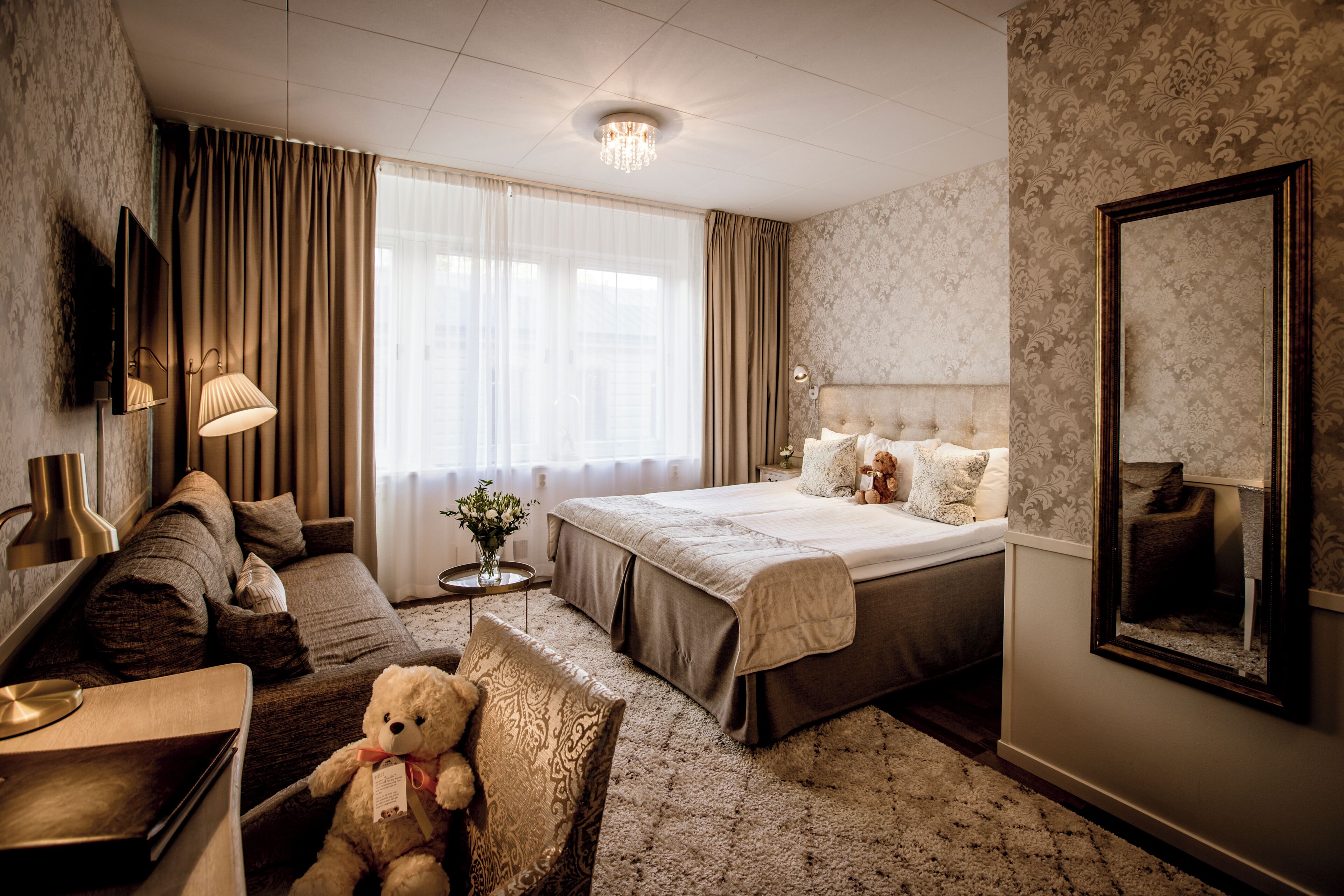 Une chambre d’hôtel avec un lit, un canapé, une chaise, un ours en peluche, une lampe, un miroir et une fenêtre avec des rideaux.