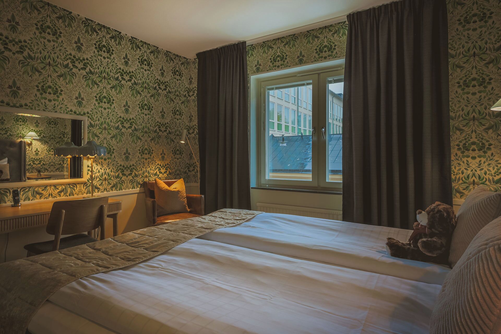 Une chambre d’hôtel avec un lit, un bureau, une chaise, une fenêtre avec des rideaux et un ours en peluche.