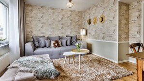 A living room with a grey sofa, a fluffy rug, a small round table, and a wall with vintage-style wallpaper. - Corner Hotel (Stockholm)