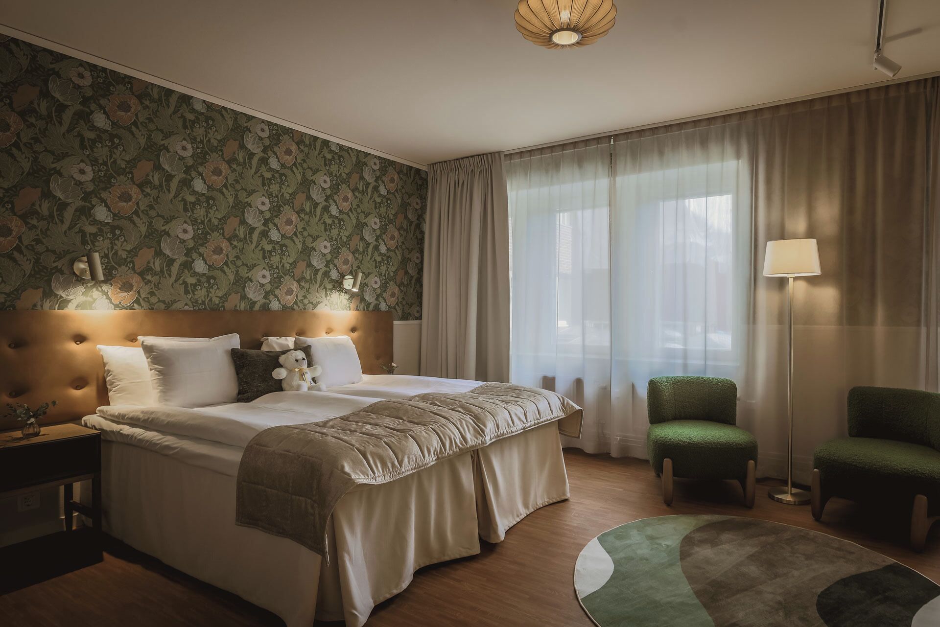 A hotel room with a large bed, two bedside tables, a green armchair, a floor lamp, and a window with sheer curtains.