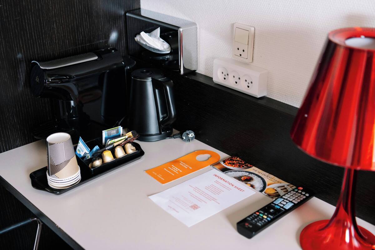 A hotel room with a coffee maker, a remote control, a red lamp, a tissue box, and a menu on the counter.