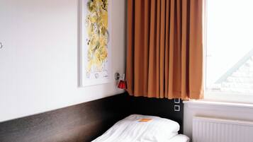 Une chambre d’hôtel avec un lit, une fenêtre avec des rideaux, un radiateur et une œuvre d’art accrochée au mur.