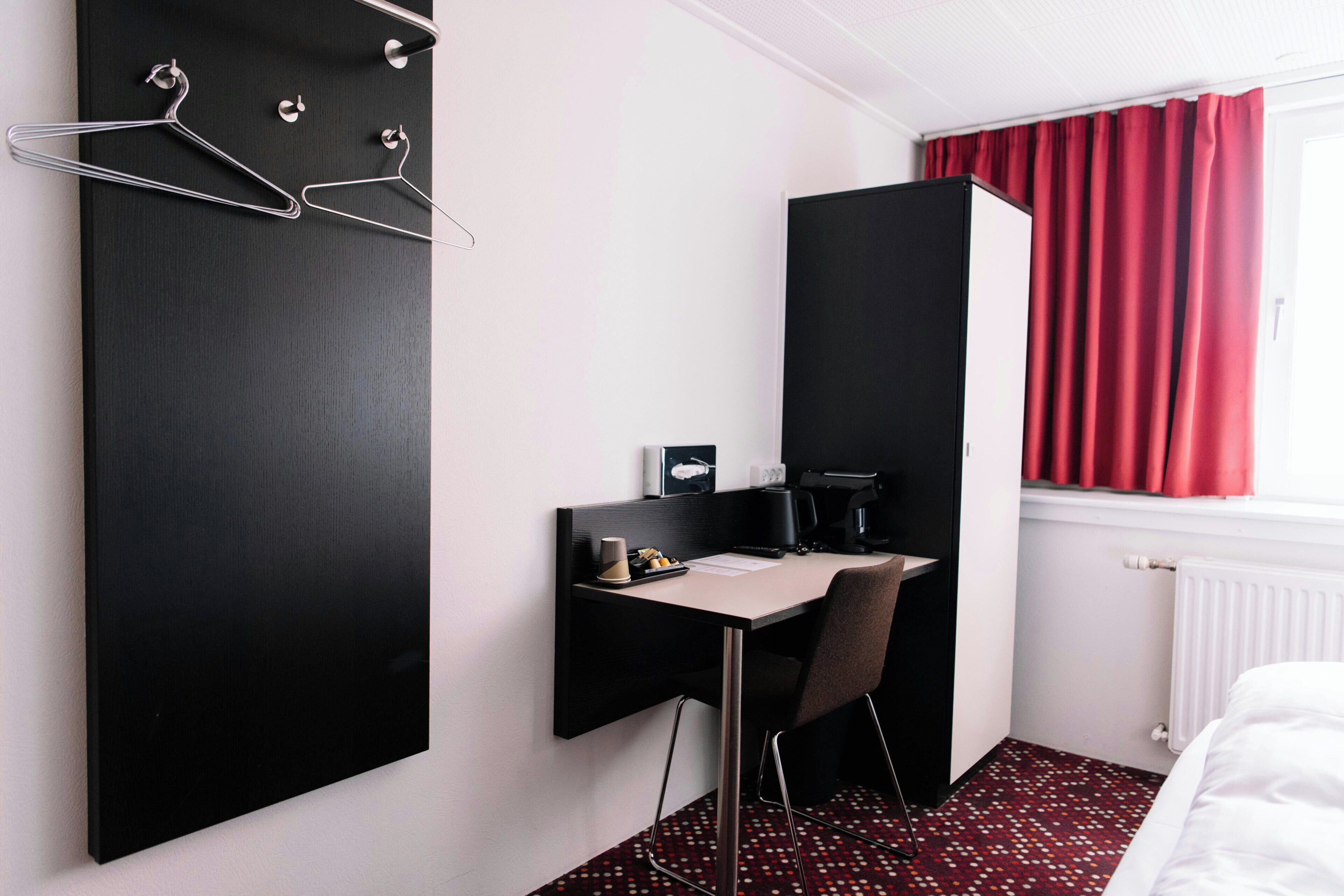 Une chambre d’hôtel avec un bureau noir, une chaise, un lit, un radiateur et une fenêtre avec des rideaux rouges.