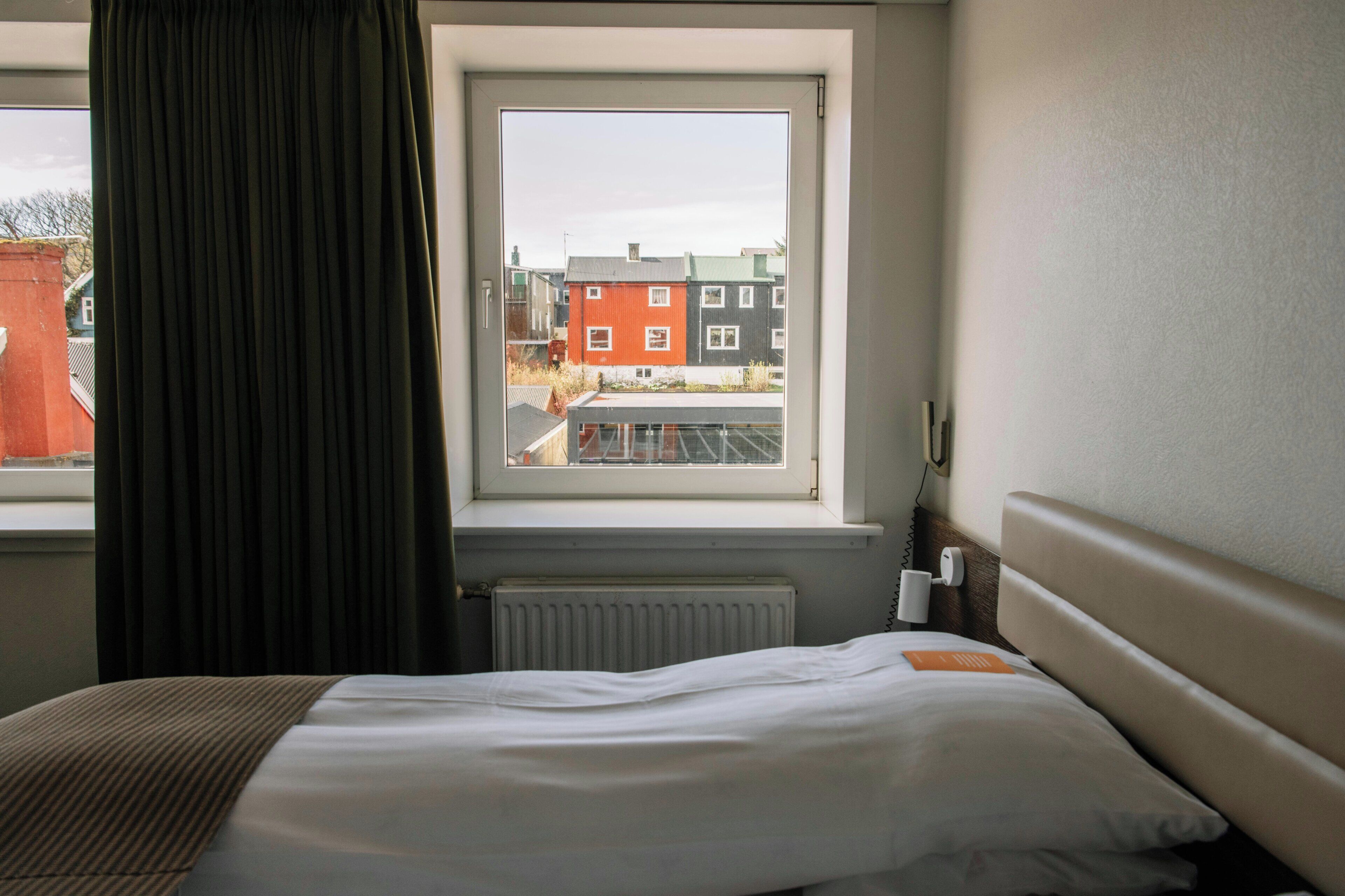 A hotel room with a bed, a window with a view of buildings, a radiator, and a bedside table with a lamp.