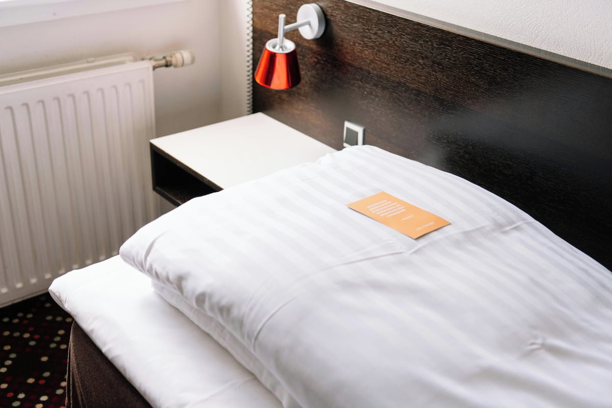 A neatly made bed with white linens, a small bedside table, a red lamp, and a white radiator.