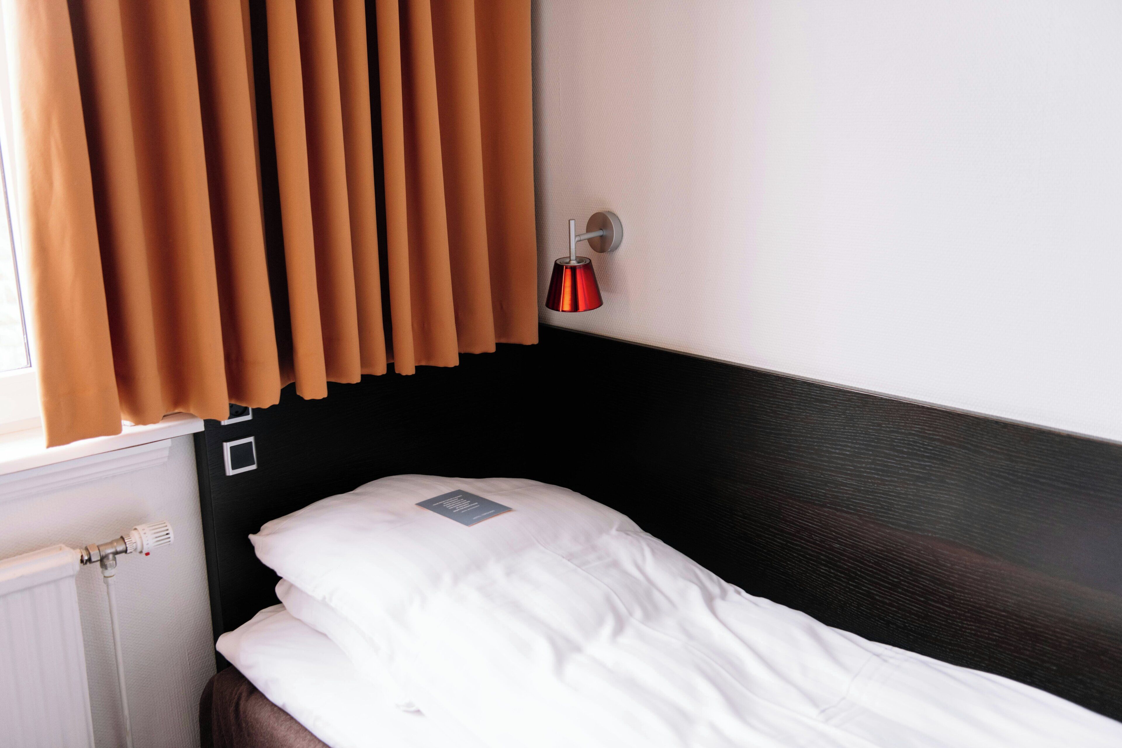 A single bed with white bedding, a wall-mounted light, and a window with brown curtains.