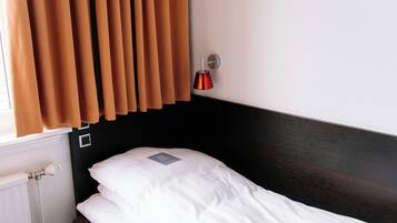 Un llit individual amb roba de llit blanca, una làmpada penjada a la paret i una finestra amb cortines marrons.