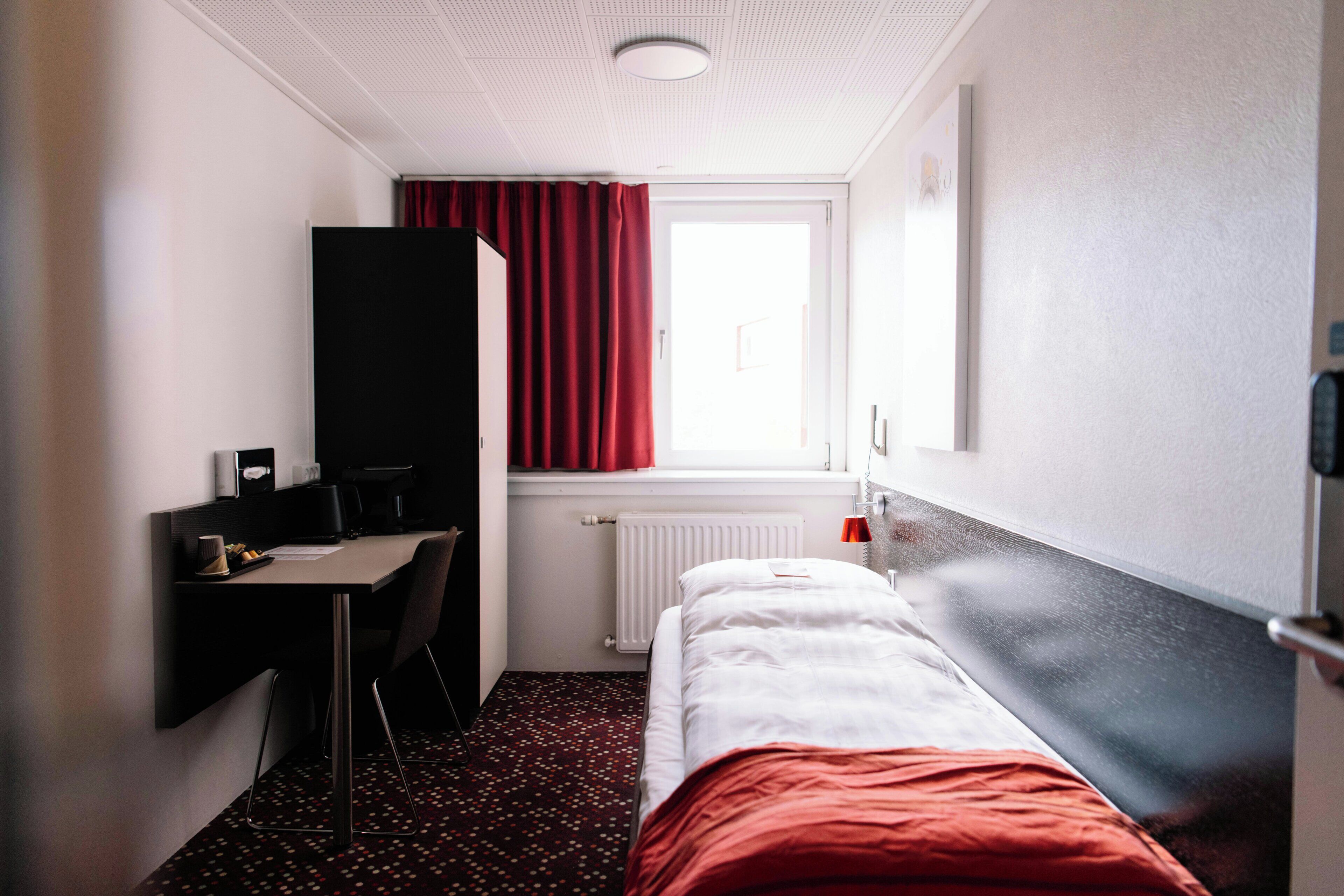 A hotel room with a bed, a desk, a chair, a radiator, and a window with curtains.
