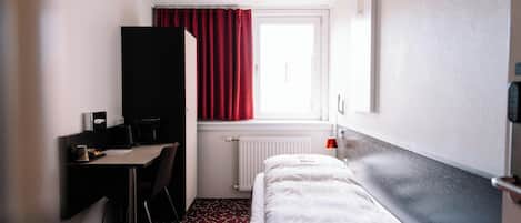 A hotel room with a bed, a desk, a chair, a radiator, and a window with curtains.