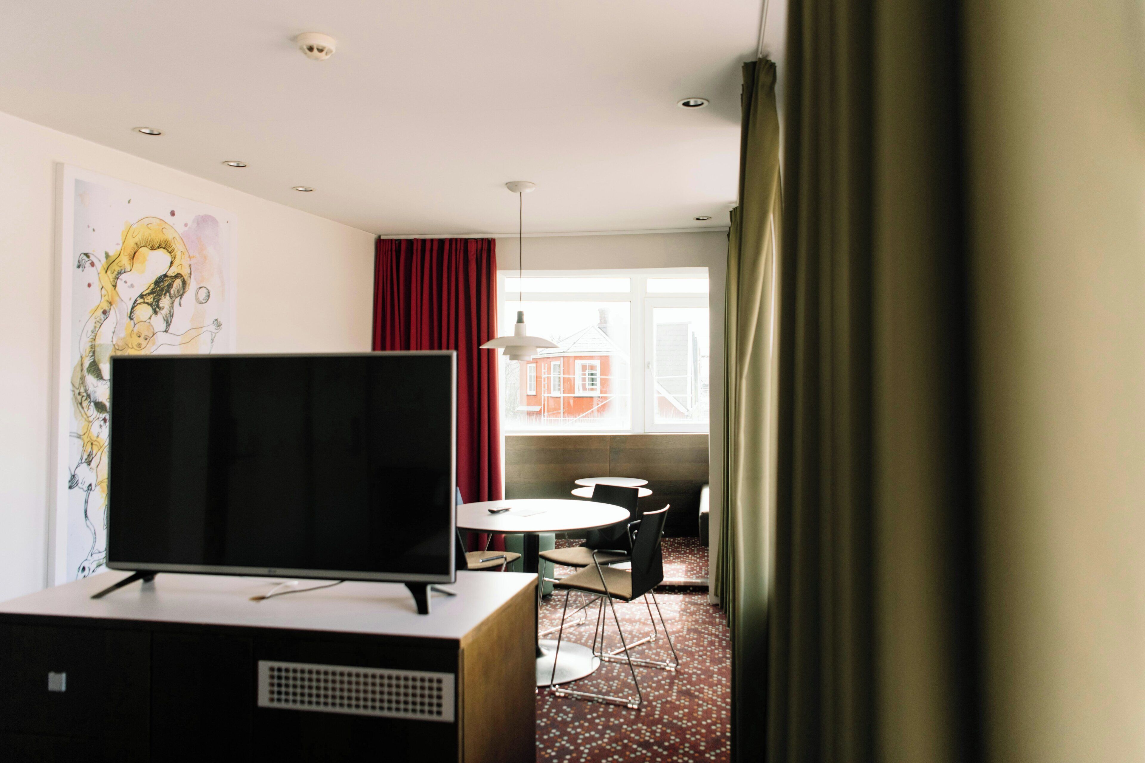 A modern hotel room with a flat-screen TV on a desk, a dining table with chairs, a large window with red curtains, and a view of buildings outside.