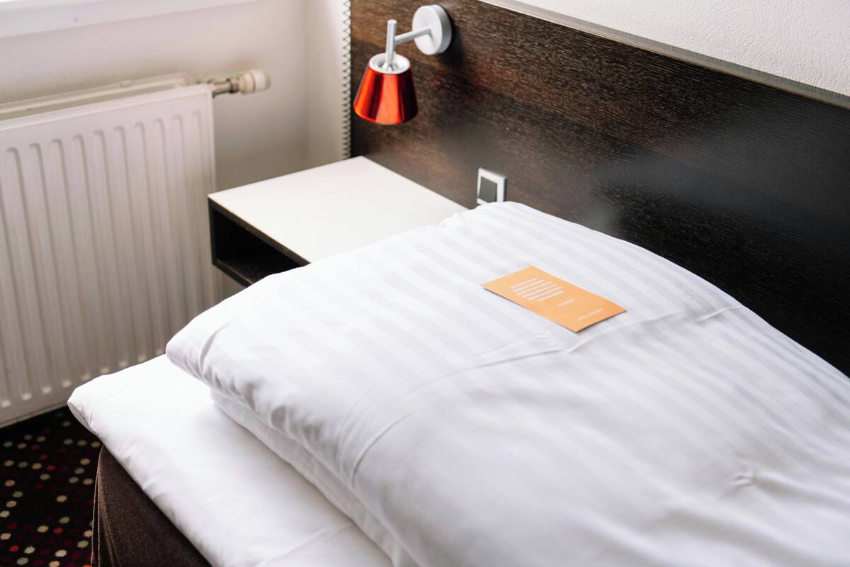 A neatly made bed with white linens, a small bedside table, a red lamp, and a white radiator.