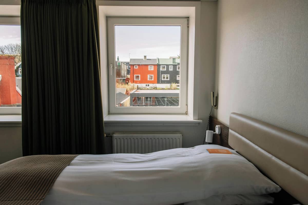 A hotel room with a bed, a window with a view of buildings, a radiator, and a bedside table with a lamp.