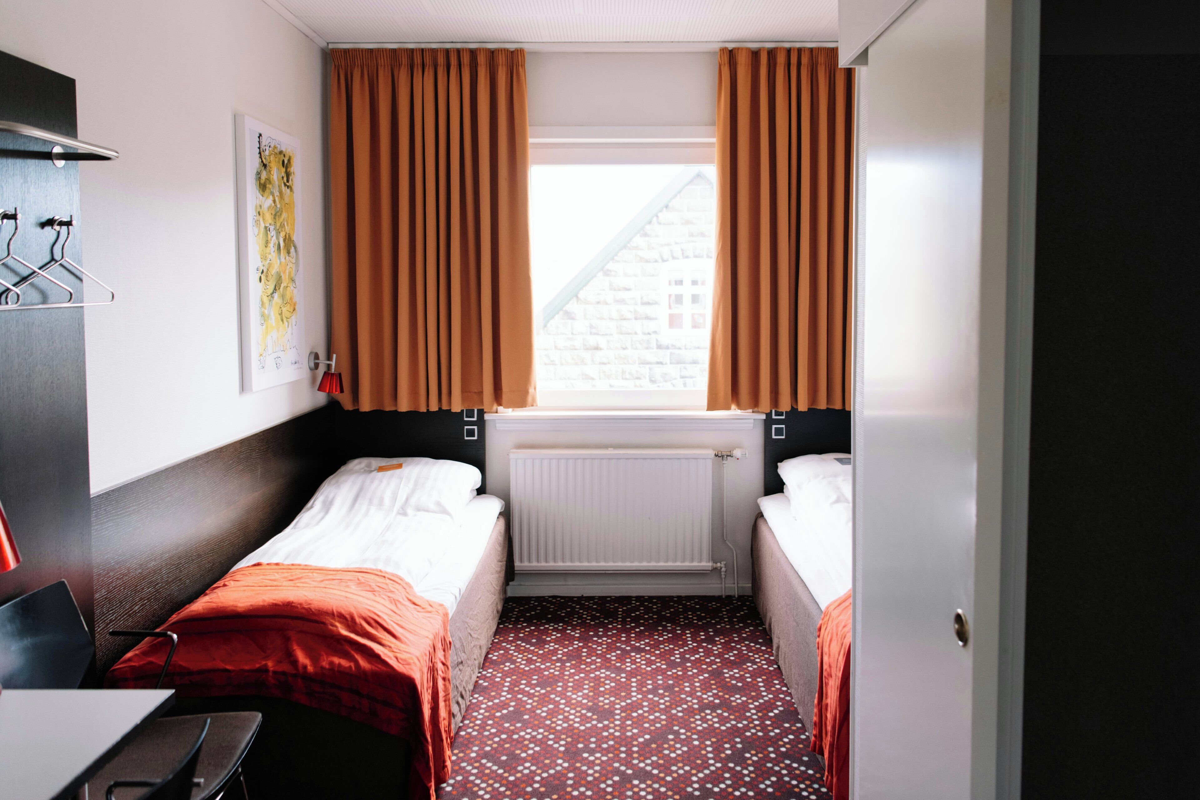 A hotel room with two beds, a radiator, a window with curtains, a desk with a chair, and a wall-mounted coat rack.