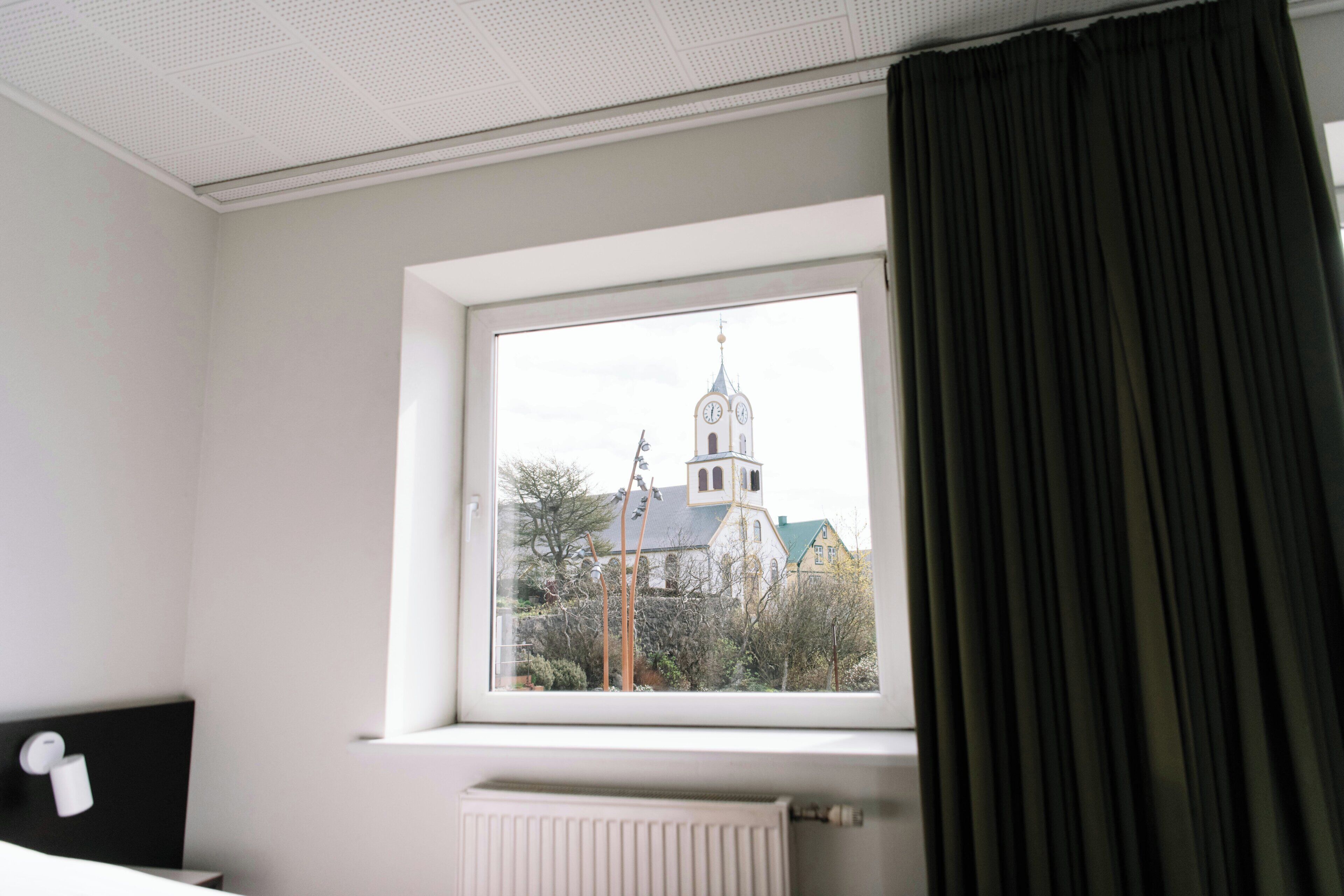A hotel room with a large window, a radiator below, and a view of a church with a clock tower.