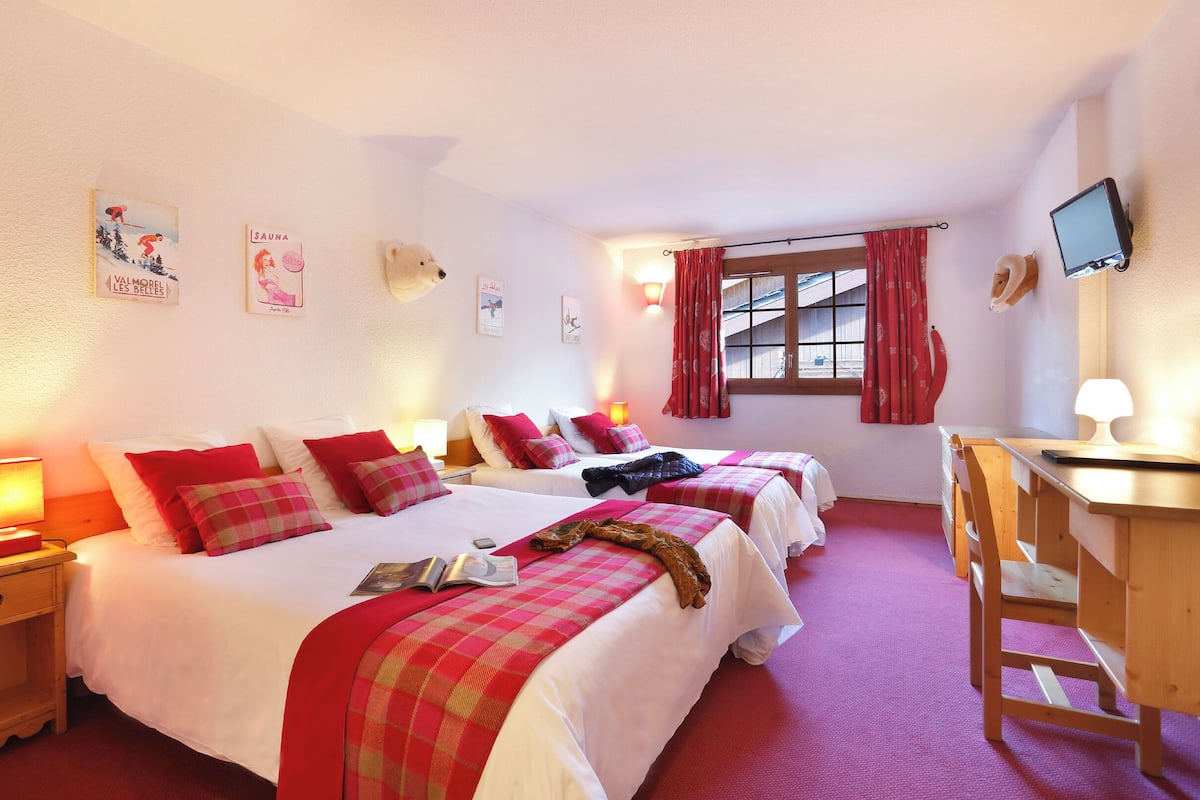 A hotel room with three beds, red and white bedding, a desk with a chair, and a TV mounted on the wall.