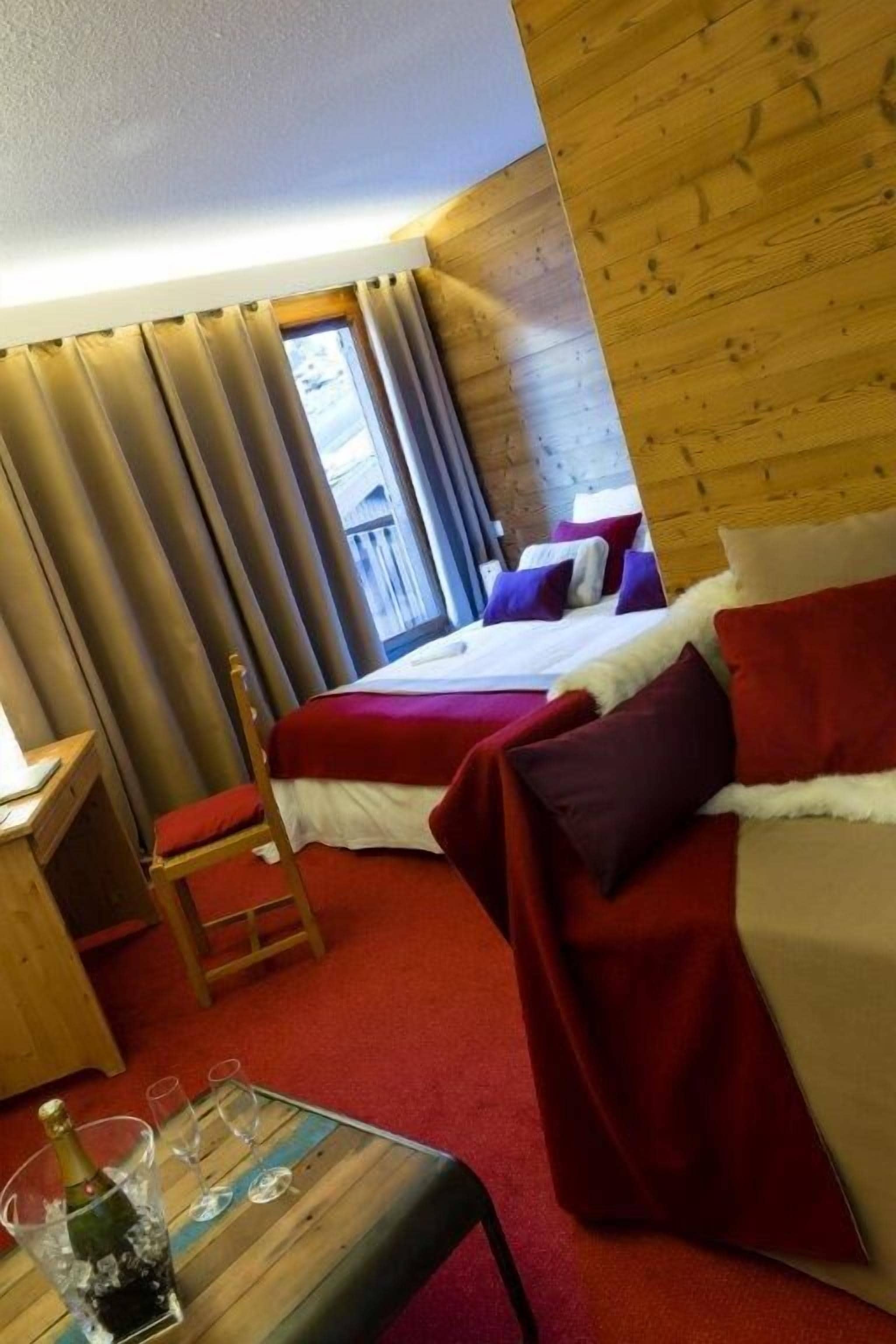 A hotel room with a wooden headboard, a bed with red and white bedding, a small wooden table with a bottle and glasses, and a view of a balcony through large curtains.