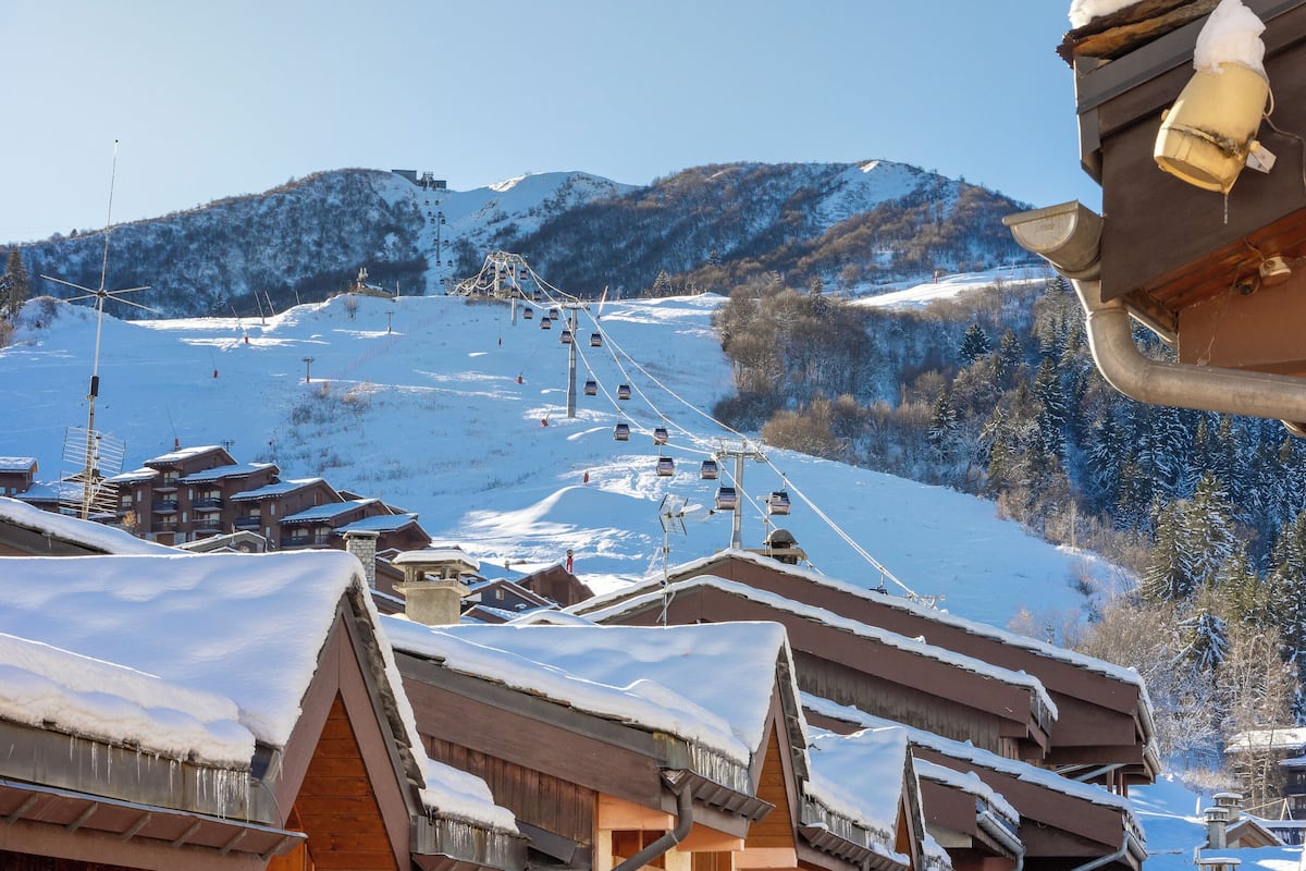 A snow-covered village with wooden buildings, a ski lift in the background, and a clear sky.