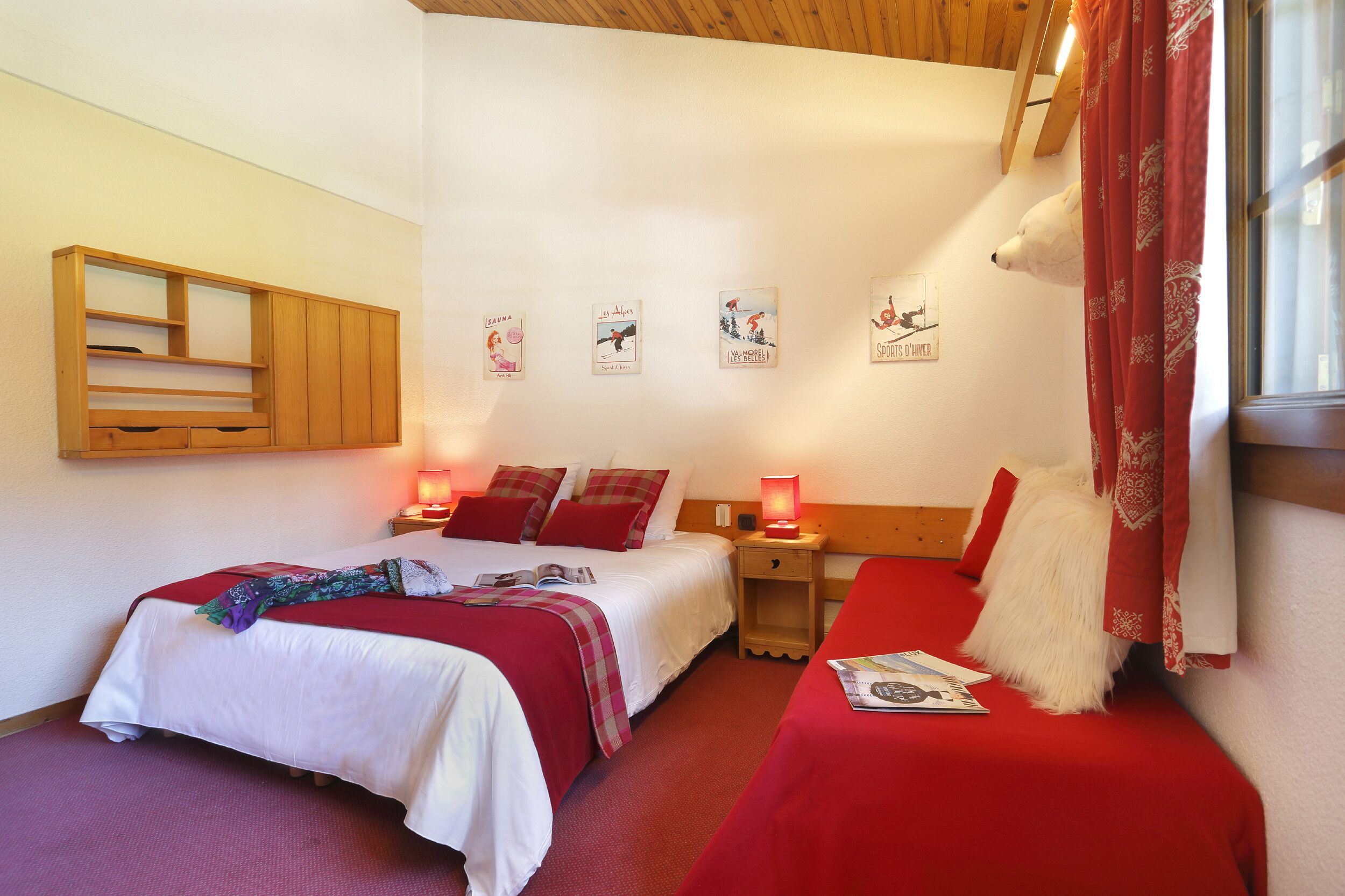 Une chambre d’hôtel avec deux lits, une tête de lit en bois, une couvre-lit rouge et blanche, un plaid rouge et blanc, un oreiller rouge et blanc, une table de chevet en bois, une lampe rouge et blanche et une fenêtre avec des rideaux rouges.