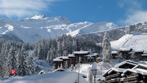 Un complejo turístico nevado con edificios de estilo chalet, cielo despejado y un pico montañoso destacado al fondo.