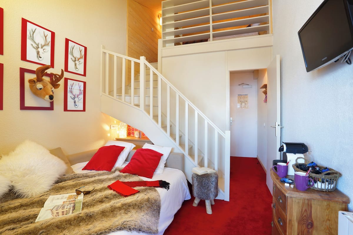 A bedroom with a bed, red pillows, a white headboard, a wooden staircase, a TV on the wall, and a red carpet.