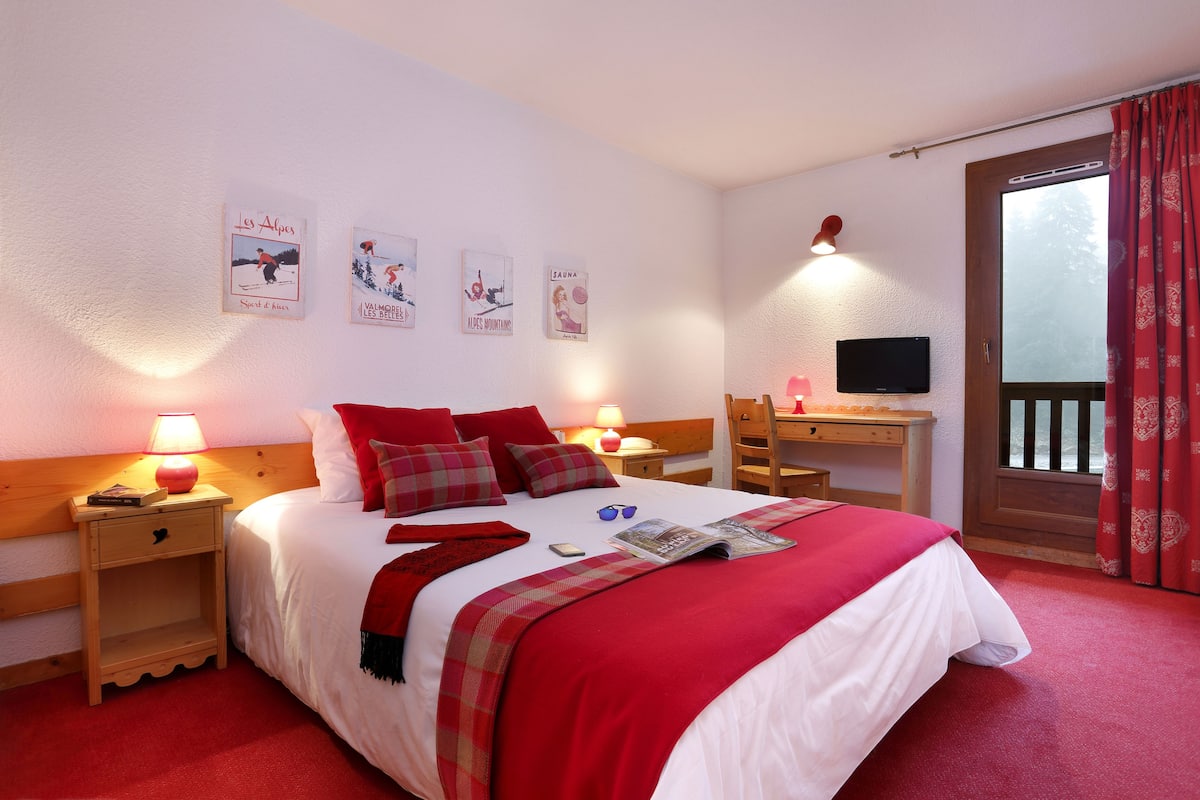 A neatly made bed with red and white bedding, a bedside table with a lamp, a desk with a computer, and a TV.