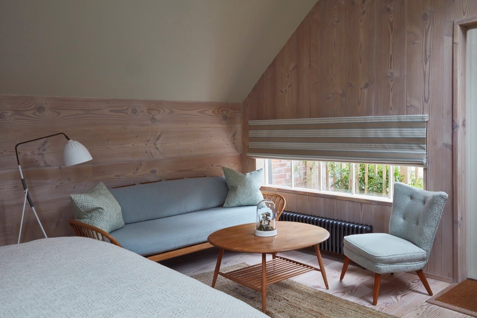 A cozy room with a wooden sloped ceiling, a light blue sofa, a round wooden table, and a single armchair.