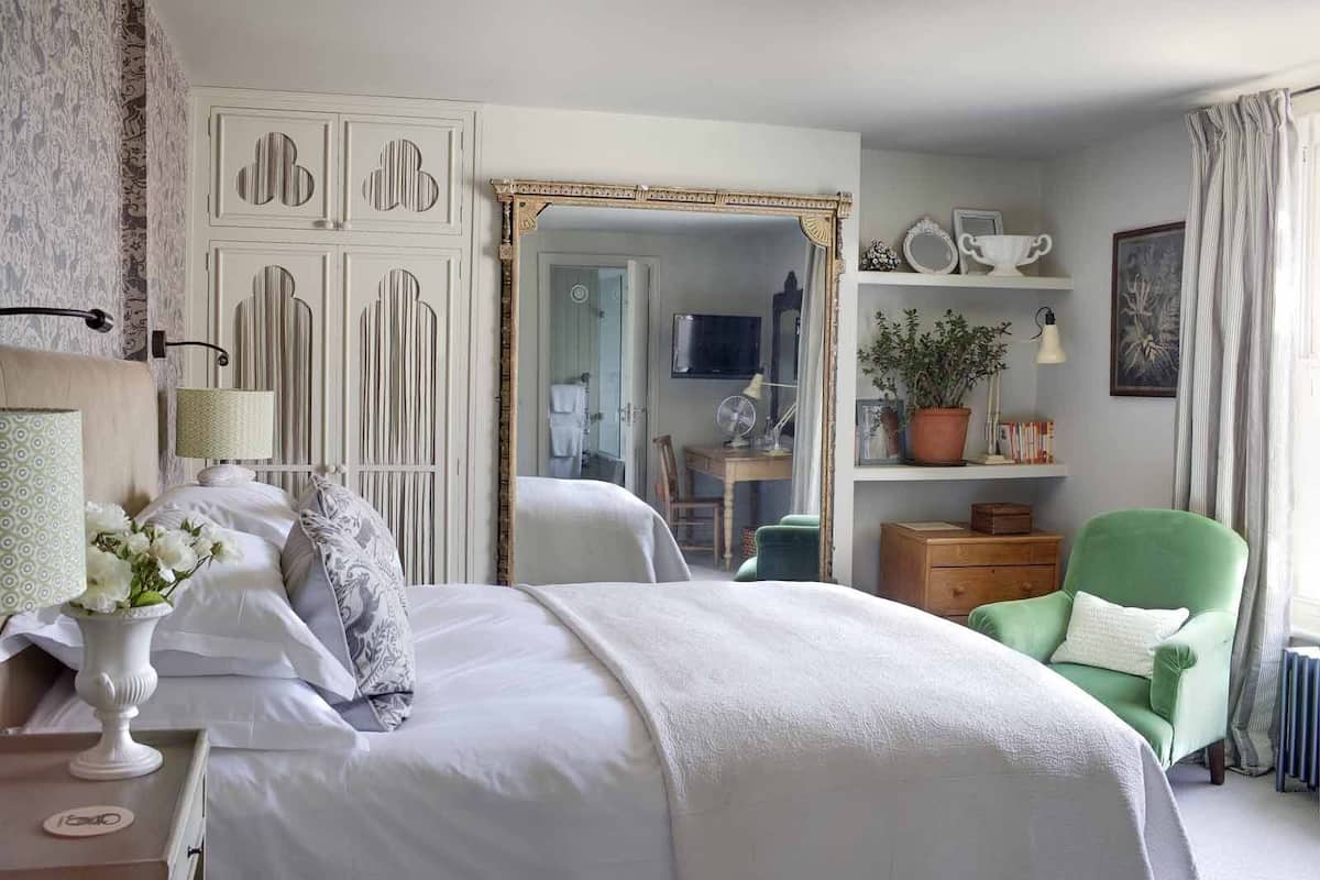 Habitación de hotel con dos camas, un sillón verde, un espejo, un escritorio con silla, una estantería y un radiador.