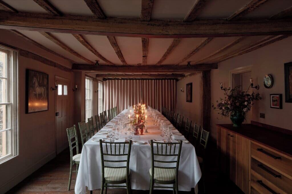 A long dining table set for a meal with chairs, wooden beams on the ceiling, and a window on the left.