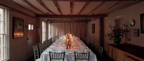 A long dining table set for a meal with chairs, wooden beams on the ceiling, and a window on the left.