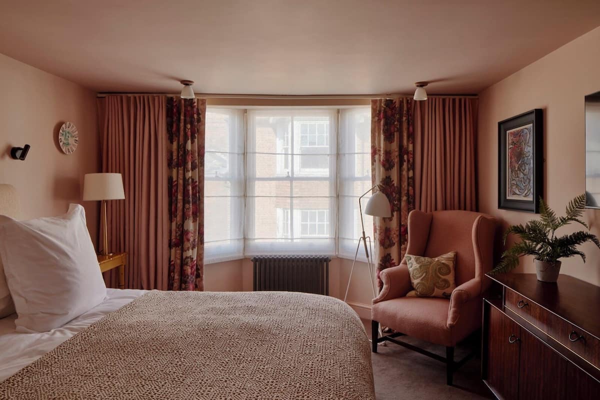 Habitación de hotel con cama, una silla, escritorio, radiador y ventana con cortinas.