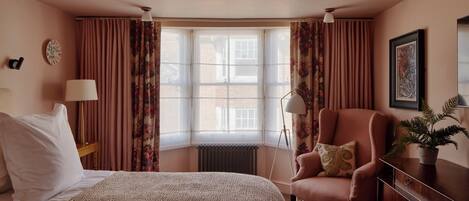 A hotel room with a bed, a chair, a desk, a radiator, and a window with curtains.